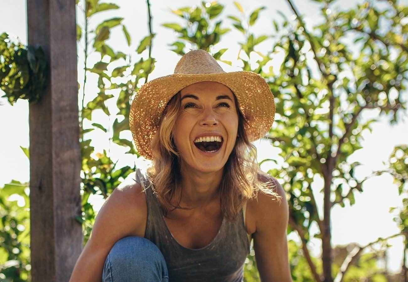 A woman wearing a straw hat and tank top smiles and laughs outdoors in a sunlit garden with leafy green trees in the background.