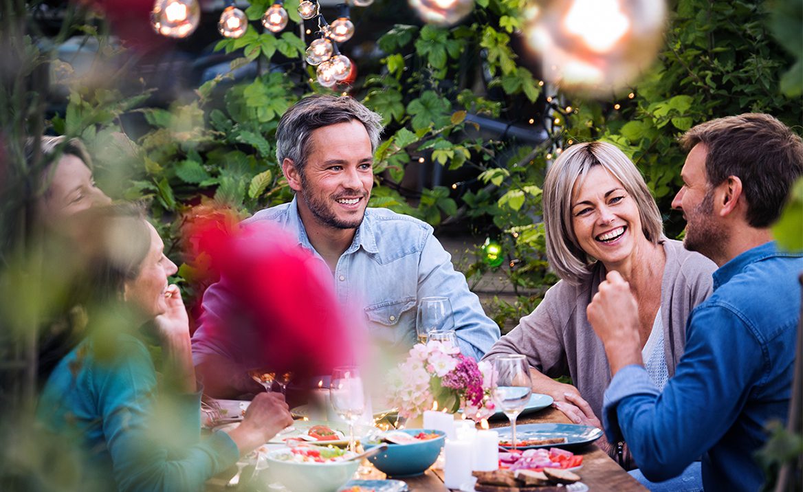Four adults sit around an outdoor table decorated with flowers and food, smiling and engaged in conversation under hanging string lights, surrounded by lush greenery.