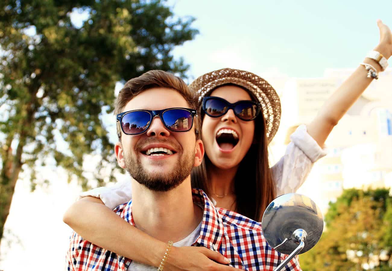 A happy young couple wearing sunglasses smiles outdoors; the woman hugs the man from behind, raising one arm in excitement. They appear joyful, with trees and buildings in the sunlit background.