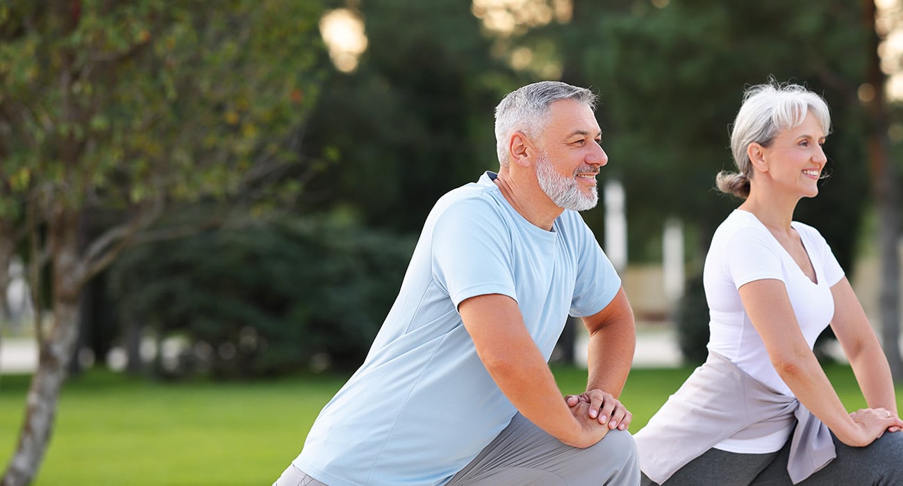 Two older adults, a man and a woman, are exercising outdoors in a park, smiling as they perform lunges on the grass. They appear healthy and active, wearing comfortable athletic clothing.